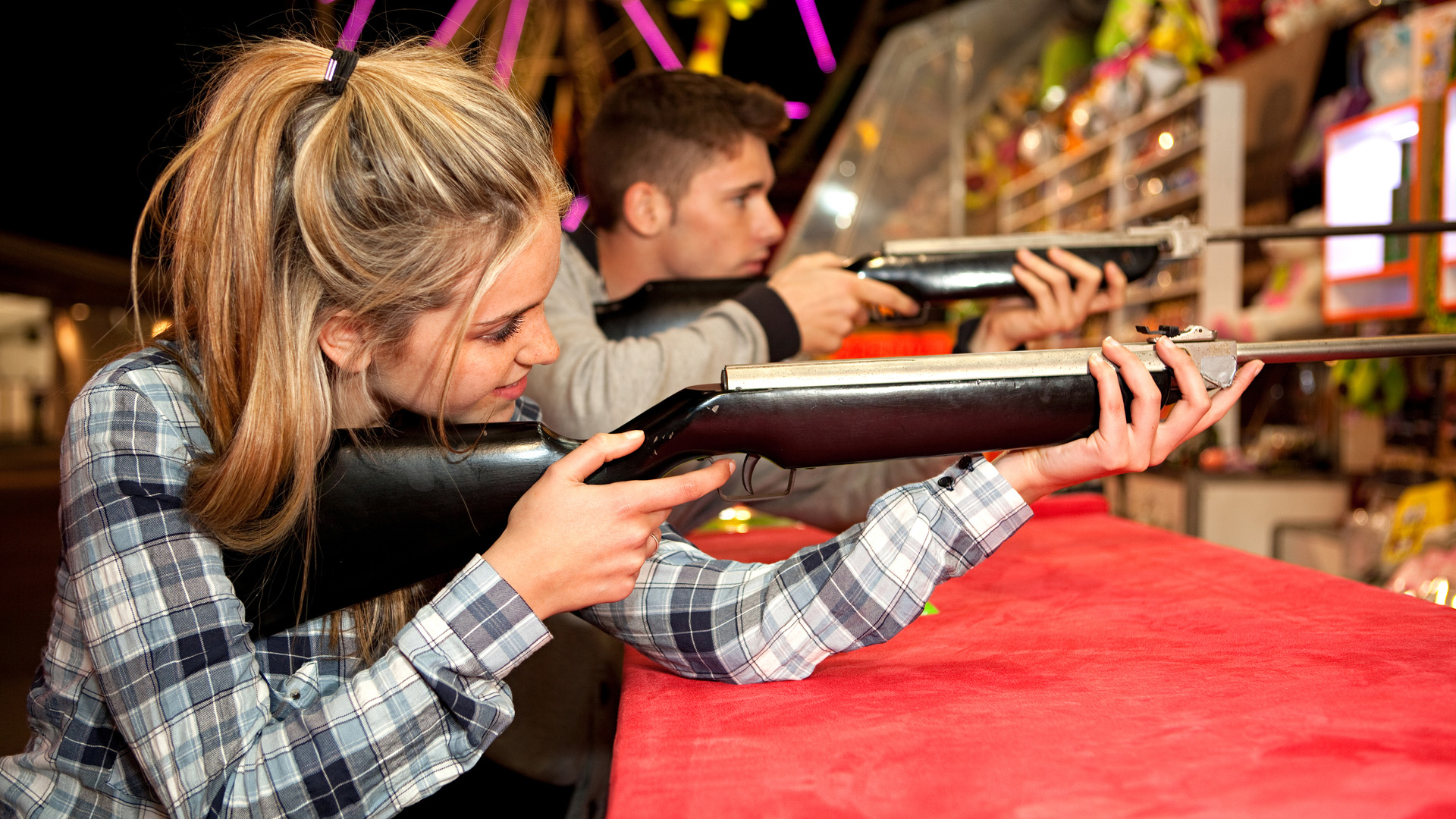 Blonde woman in a checkered shirt and man in a jacket, both holding rifles and taking aim at an amusement park.
