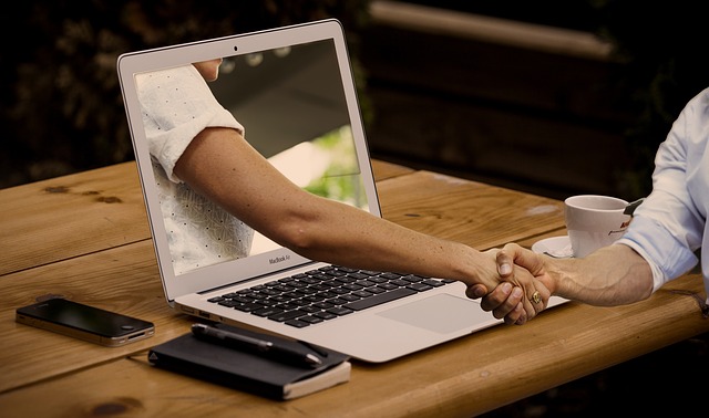 A person sitting at a desk with coffee, mobile, notepad, pen and laptop shaking another persons hand through the screen